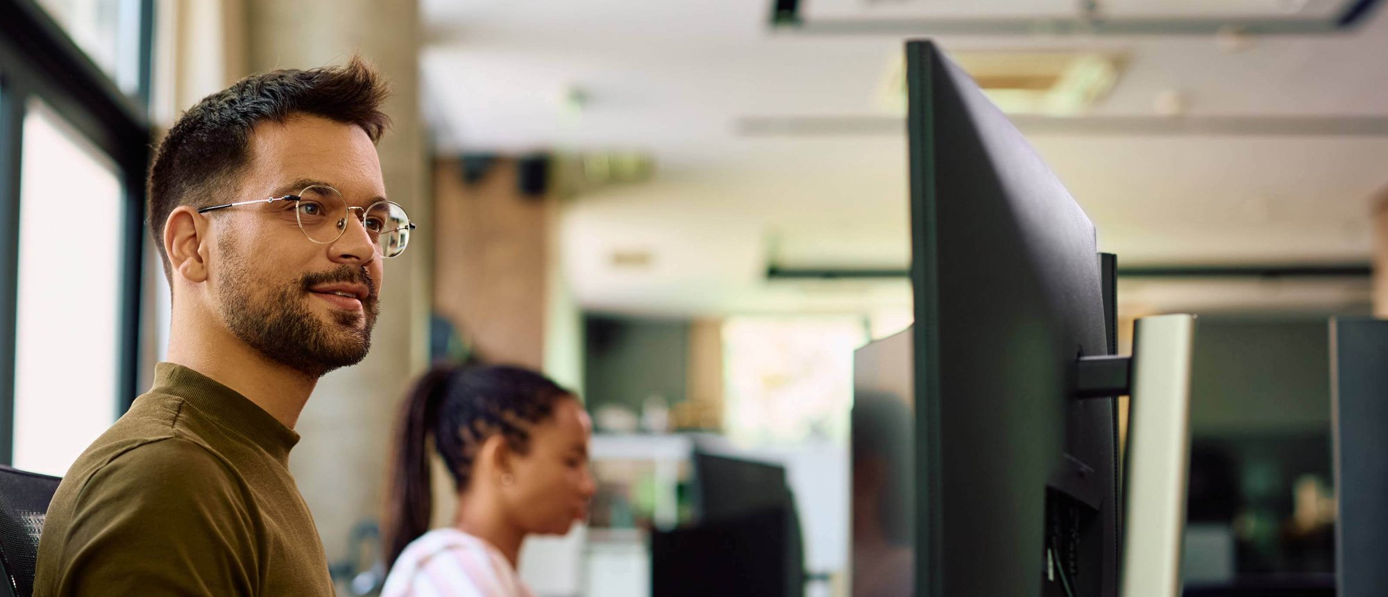 Man with glasses looking at computer screen with woman in background