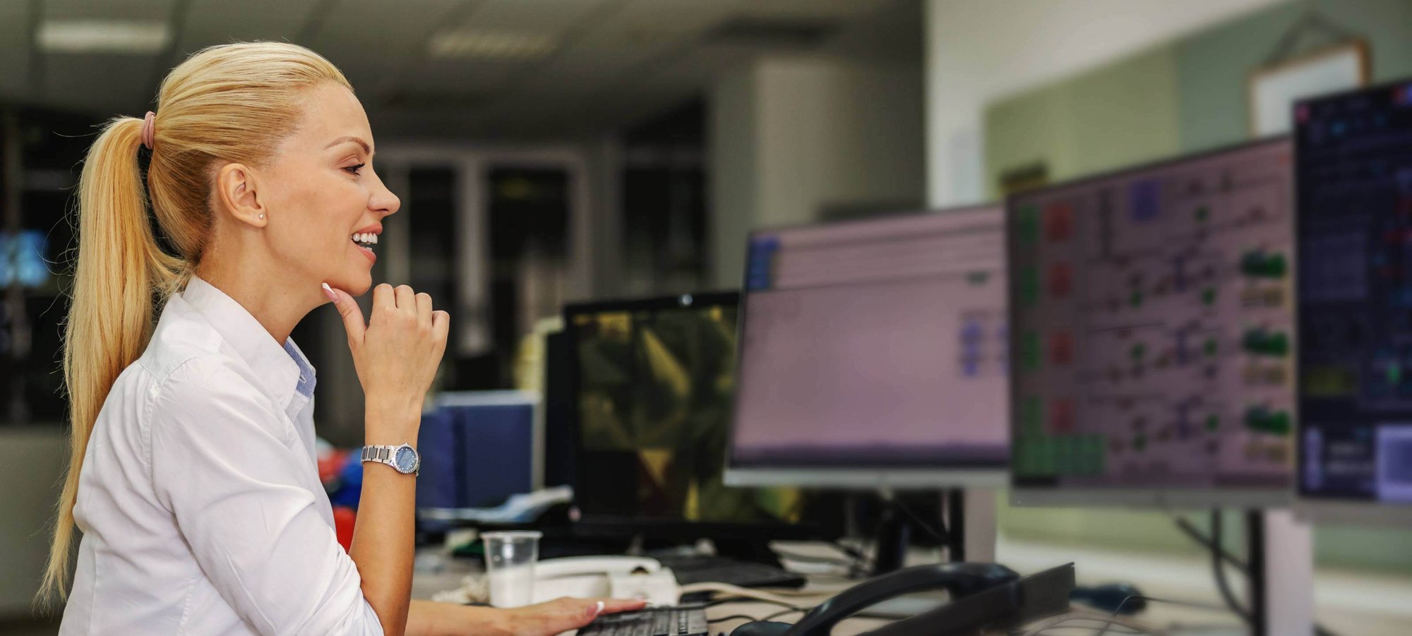 Woman in shirt smiling with thumb on chin looking at computer monitors